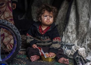 A displaced Palestinian child eats food from a box while sitting in a makeshift tent at a camp beside a street in Rafah / ©AFP