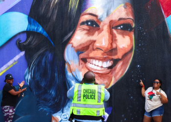 A Chicago police officer photographs people posing before a mural of US Vice President Kamala Harris outside the United Center ahead of the Democratic National Convention / ©AFP